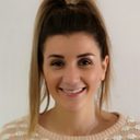 Portrait of a person with a high ponytail, smiling, in a cream spotted jumper against a pale wall.