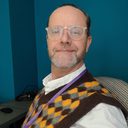 Portrait of JD, with glasses and a beard, in a brown and yellow argyle tank top with a lanyard, against a teal wall.