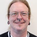Portrait of a person with long hair tied back, glasses, ear plugs and facial piercings, in a black cardigan over a white t-shirt.