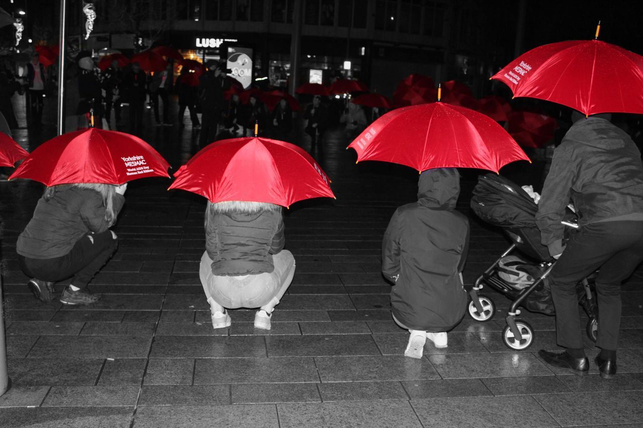 Selective-colour night photo of people crouching under red Yorkshire MESMAC umbrellas on a wet pavement, one with a pram, marking World AIDS Day.