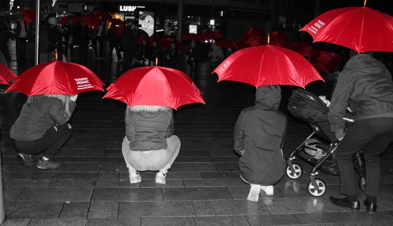 Selective-colour night photo of people crouching under red Yorkshire MESMAC umbrellas on a wet pavement, one with a pram, marking World AIDS Day.