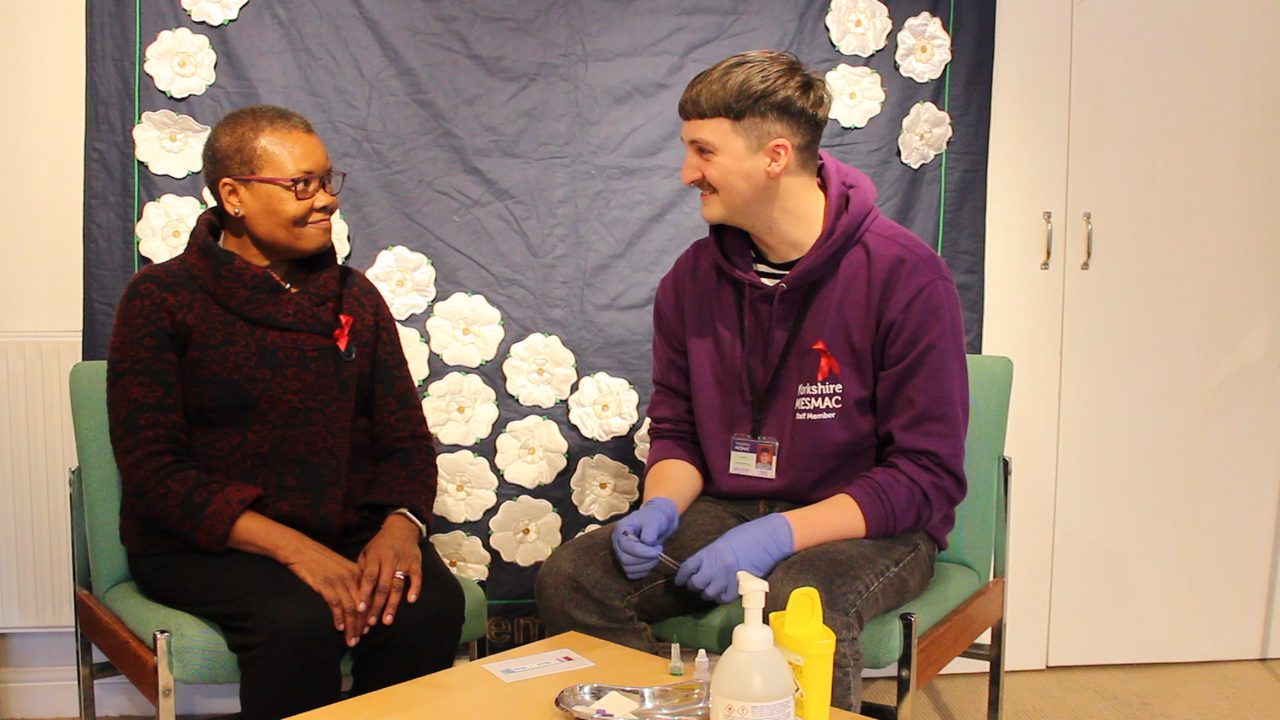A person in glasses wearing a red ribbon sitting and smiling beside a Yorkshire MESMAC staff member in a purple hoodie and gloves, at an HIV testing session with a white-rose backdrop.