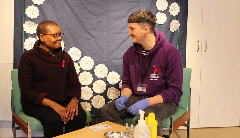 A person in glasses wearing a red ribbon sitting and smiling beside a Yorkshire MESMAC staff member in a purple hoodie and gloves, at an HIV testing session with a white-rose backdrop.