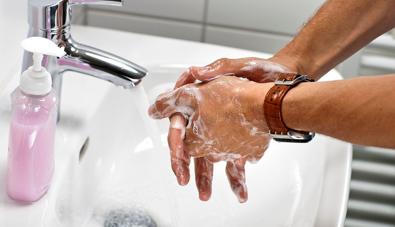 Close-up of soapy hands being washed under a running tap, beside a bottle of pink liquid soap.