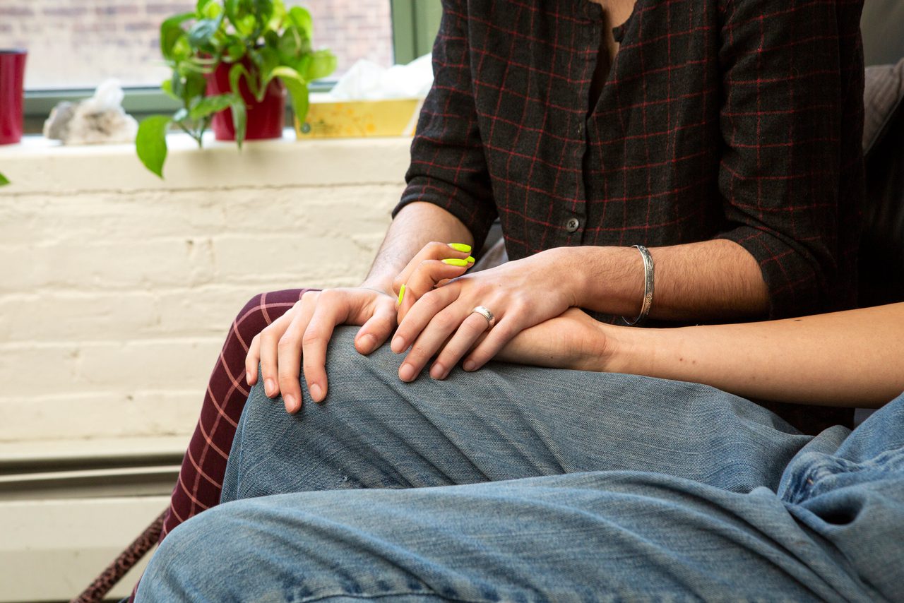 Close-up of two people sitting side by side holding hands on their laps, one with neon-painted nails, beside a window with a plant.