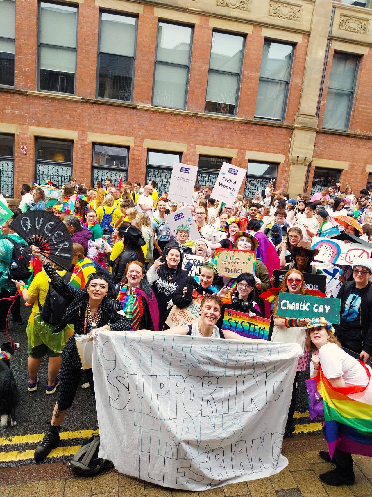 A photo of a crowd of MESMAC staff and volunteers are Leeds Pride. They are waving various banners, placards, fans and signs