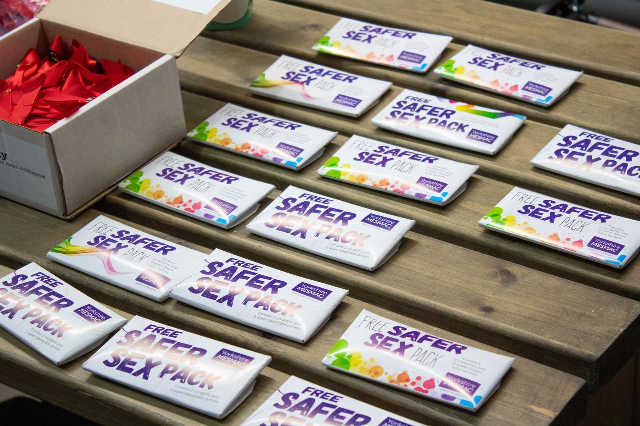 Yorkshire MESMAC "Free Safer Sex Pack" boxes laid out in rows on a wooden bench, beside a box of red ribbons.