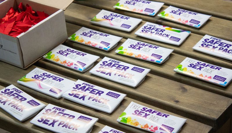 Yorkshire MESMAC "Free Safer Sex Pack" boxes laid out in rows on a wooden bench, beside a box of red ribbons.