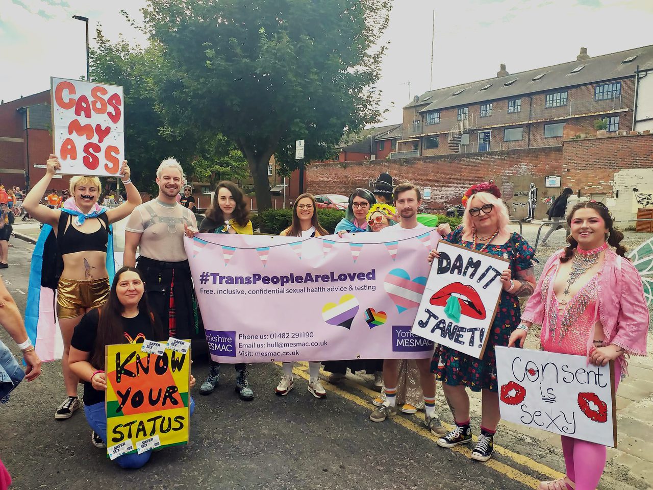 MESMAC staff and volunteers at Hull Pride, stood around a banner that reads #TransPeopleAreLoved and holding placards which say things like "Know your status", "Dam it Janet" and "Consent is sexy"