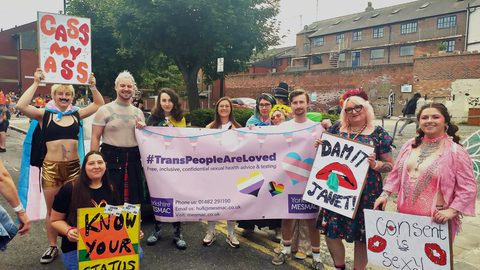 MESMAC staff and volunteers at Hull Pride, stood around a banner that reads #TransPeopleAreLoved and holding placards which say things like "Know your status", "Dam it Janet" and "Consent is sexy"