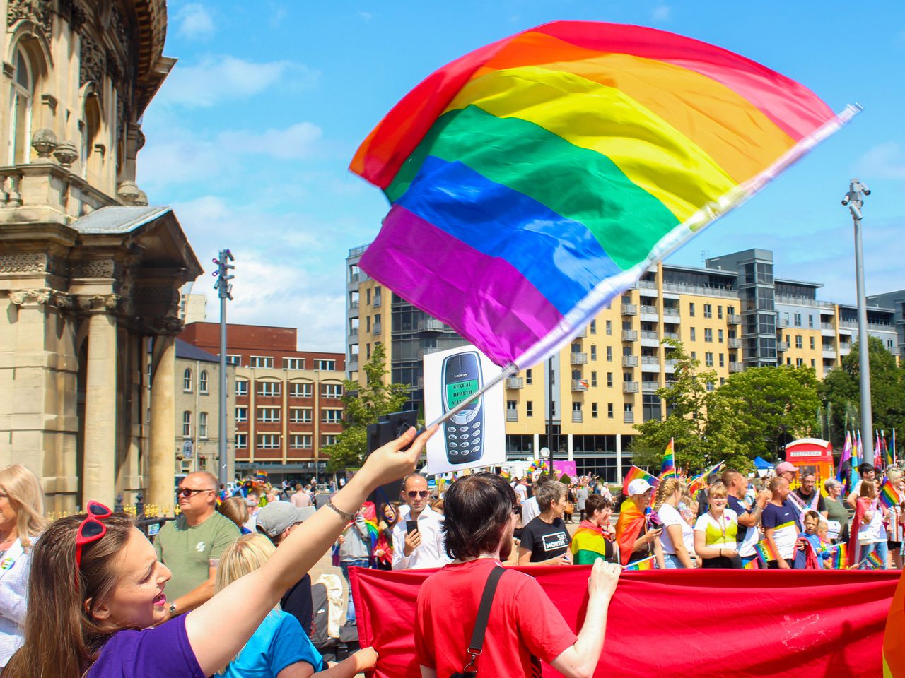 A person waves a Pride flag at Pride in Hull in Queen's Square. Behind them is a crowd carrying banners, placards and flags