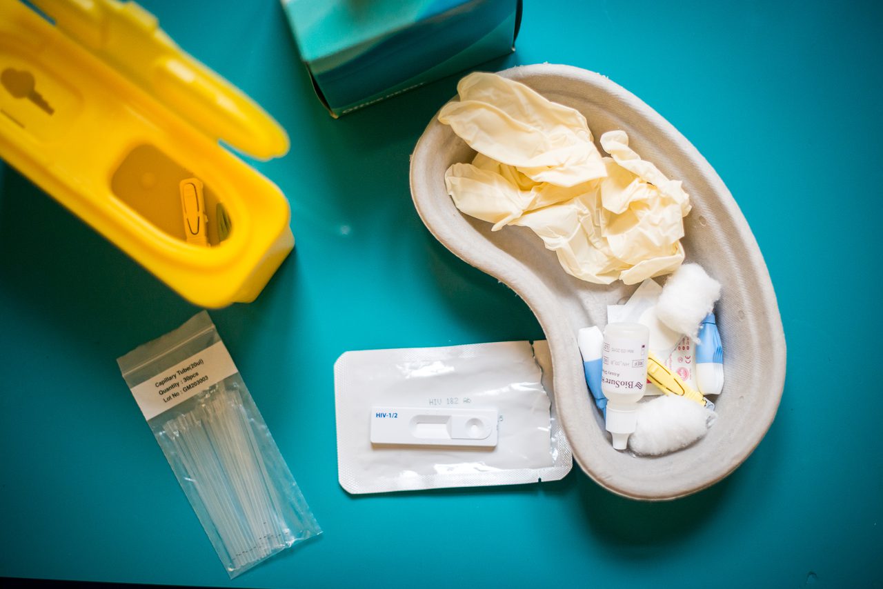 An HIV self-test kit on a teal surface: a yellow sharps bin, a packet of capillary tubes, a BioSure HIV test cassette and a kidney dish holding gloves, swabs and a sample bottle.