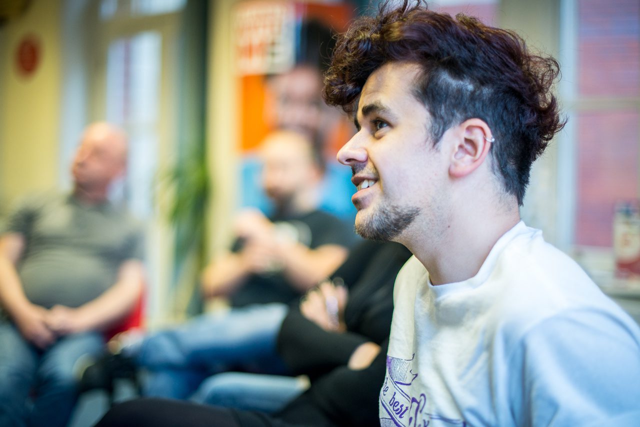 Profile of a person with curly dark hair in a white Yorkshire MESMAC t-shirt at a group session, with others blurred behind.