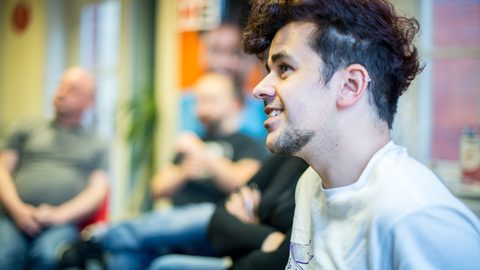 Profile of a person with curly dark hair in a white Yorkshire MESMAC t-shirt at a group session, with others blurred behind.