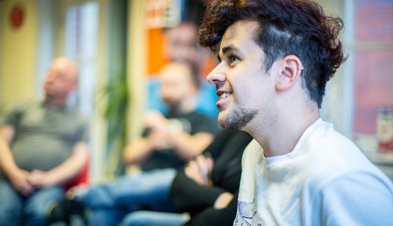 Profile of a person with curly dark hair in a white Yorkshire MESMAC t-shirt at a group session, with others blurred behind.