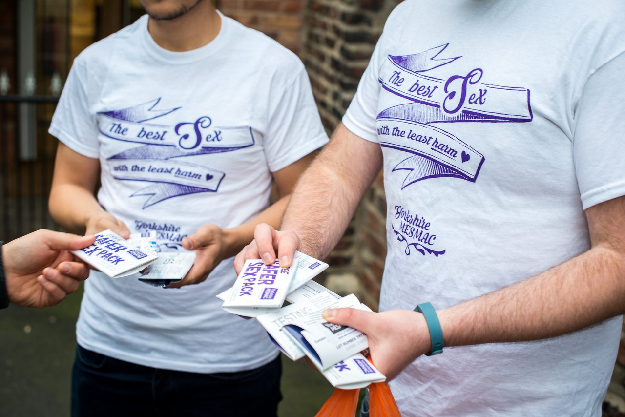 Close-up of two people in "The best sex with the least harm" t-shirts holding safer-sex packs and passing one to another hand.