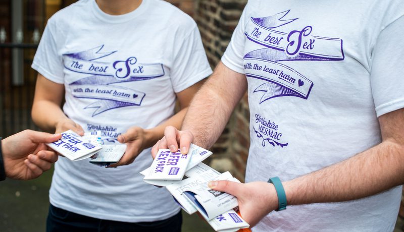 Close-up of two people in "The best sex with the least harm" t-shirts holding safer-sex packs and passing one to another hand.