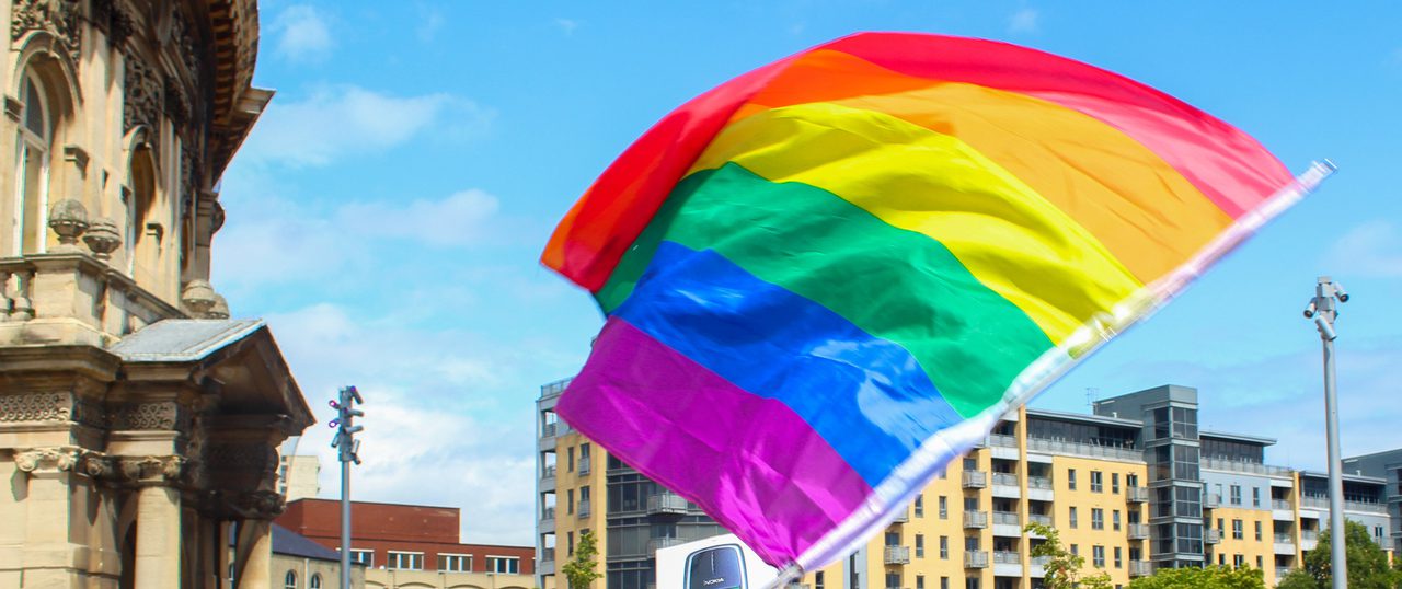 A large rainbow Pride flag waving in front of a Victorian building and modern apartments under a blue sky.