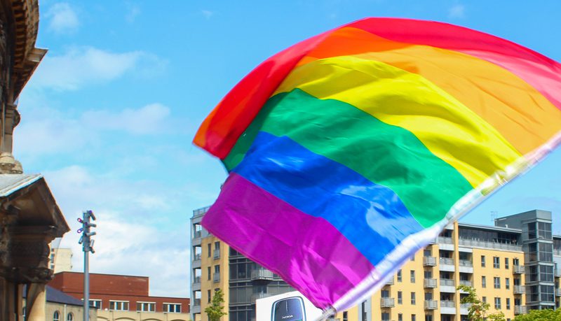 A large rainbow Pride flag waving in front of a Victorian building and modern apartments under a blue sky.