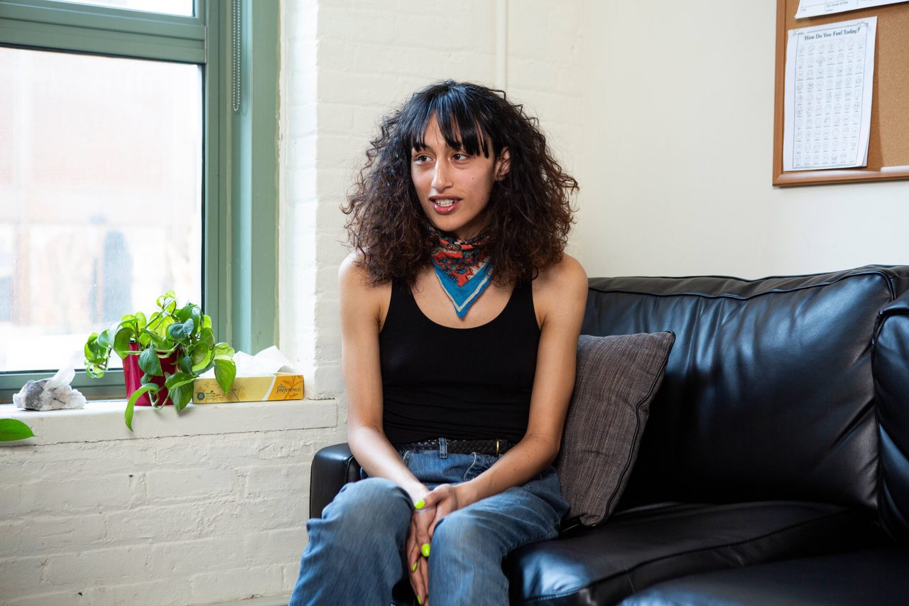A person with curly dark hair and a red neckerchief sitting on a black sofa in a black vest top, speaking to someone out of shot.