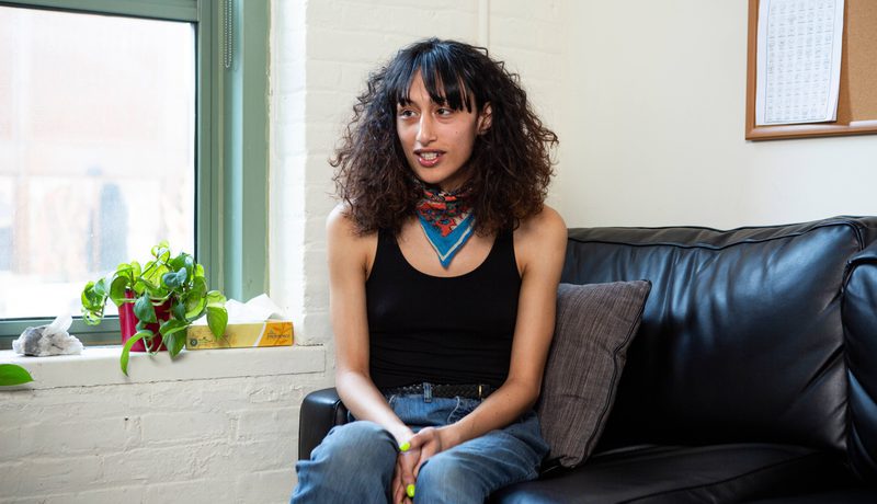 A person with curly dark hair and a red neckerchief sitting on a black sofa in a black vest top, speaking to someone out of shot.
