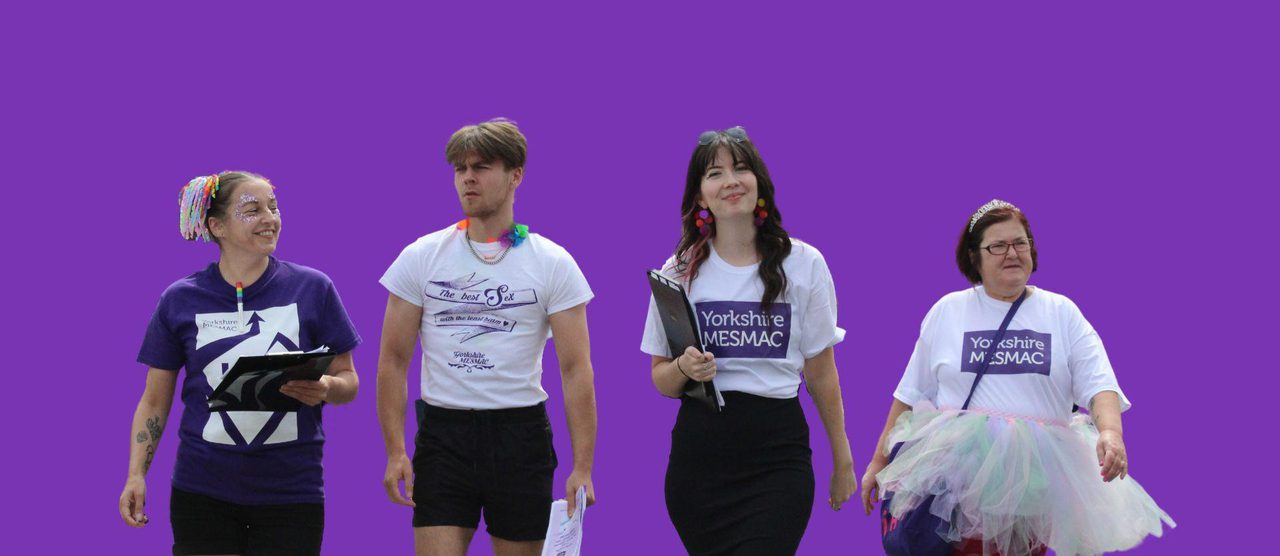 Four people walking together in Yorkshire MESMAC t-shirts against a purple background, some wearing Pride accessories and a tutu.
