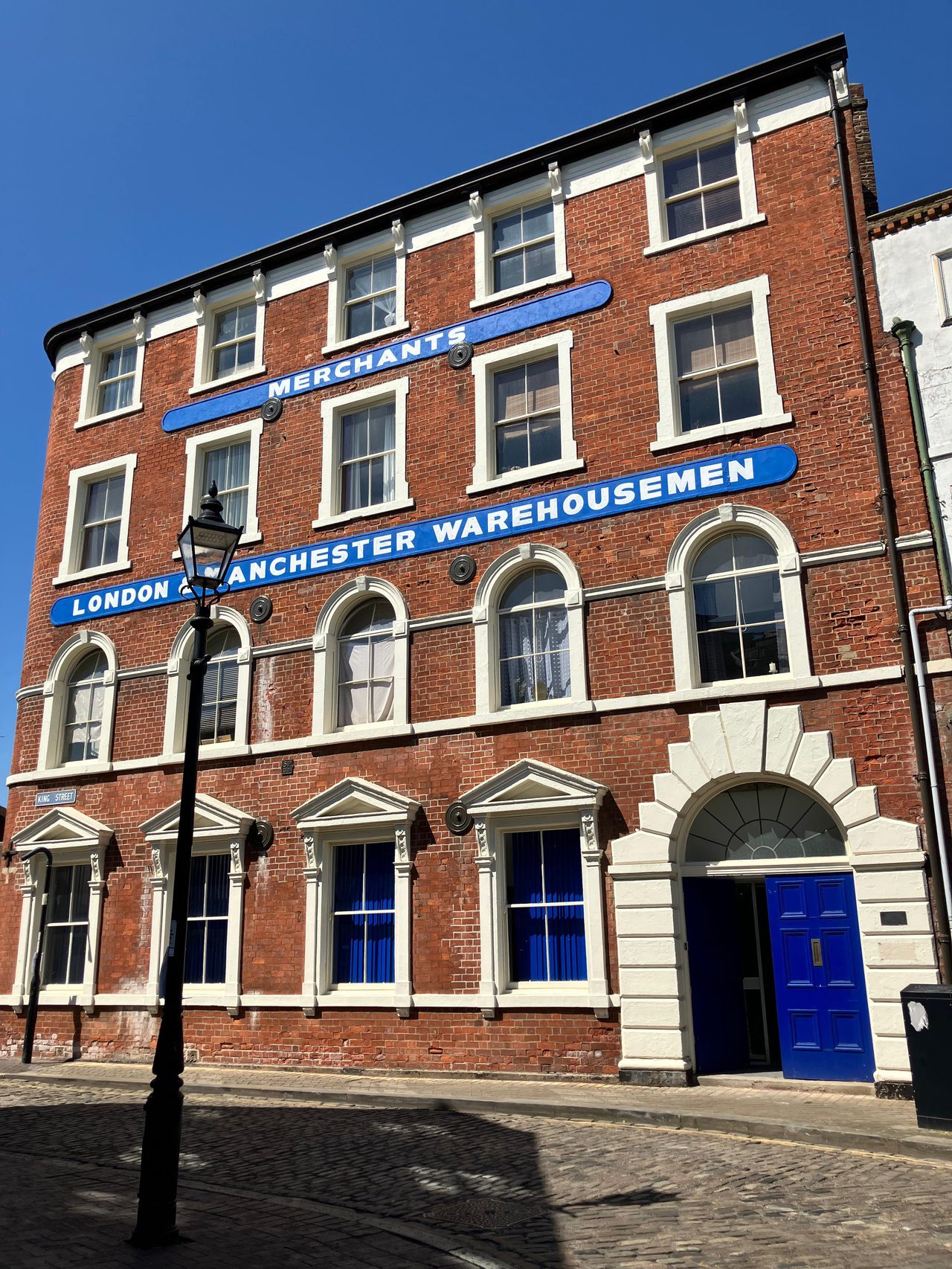 The red-brick "London & Manchester Warehousemen" building in Hull's Trinity Square, three storeys high with arched windows and a blue door standing ajar, under a blue sky.