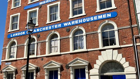 The red-brick "London & Manchester Warehousemen" building in Hull's Trinity Square, three storeys high with arched windows and a blue door standing ajar, under a blue sky.
