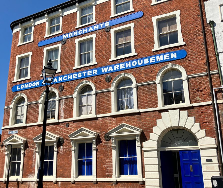 The red-brick "London & Manchester Warehousemen" building in Hull's Trinity Square, three storeys high with arched windows and a blue door standing ajar, under a blue sky.