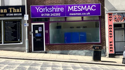 The Yorkshire MESMAC Rotherham office shopfront, a brick building with a purple "Yorkshire MESMAC" sign and a numbered door, on a sloping street beside a roast-pork shop.