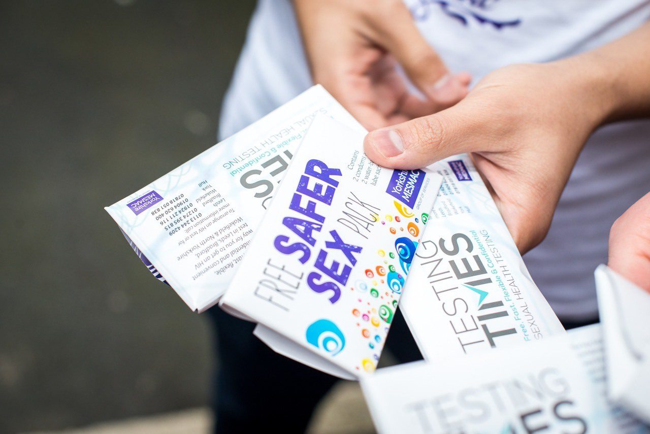 Close-up of hands holding Yorkshire MESMAC "Free Safer Sex Pack" and "Testing Times" leaflets outdoors.