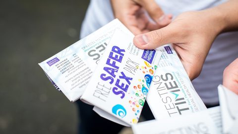 Close-up of hands holding Yorkshire MESMAC "Free Safer Sex Pack" and "Testing Times" leaflets outdoors.