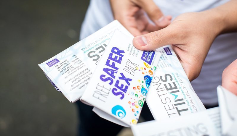 Close-up of hands holding Yorkshire MESMAC "Free Safer Sex Pack" and "Testing Times" leaflets outdoors.