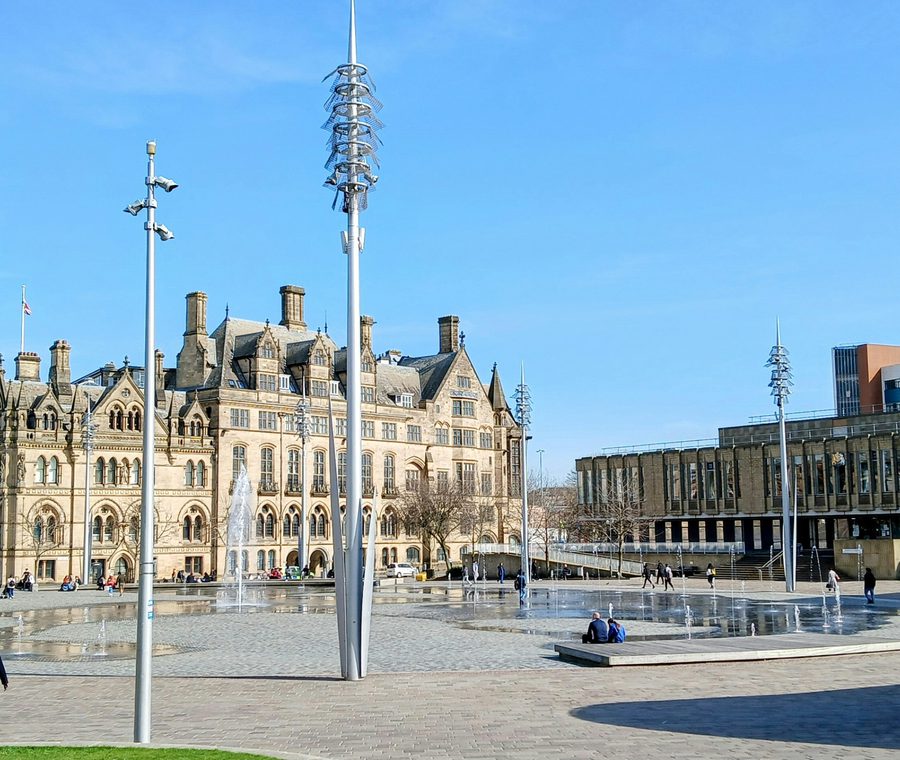 View across the Mirror Pool in Bradford City Park with people walking in the foreground and Bradford City Hall in the background under a blue sky.