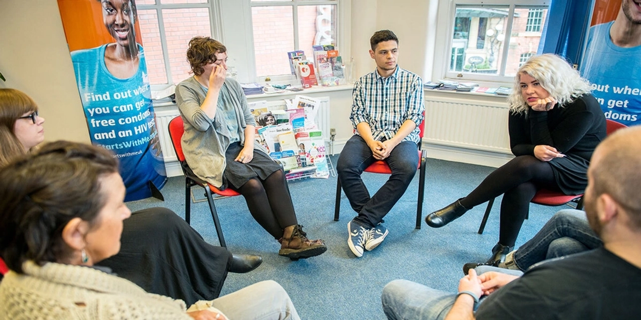 A small group sitting in a circle of chairs in a meeting room with MESMAC HIV-testing banners, talking together.