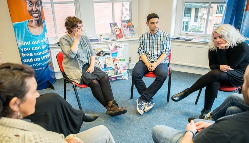 A small group sitting in a circle of chairs in a meeting room with MESMAC HIV-testing banners, talking together.
