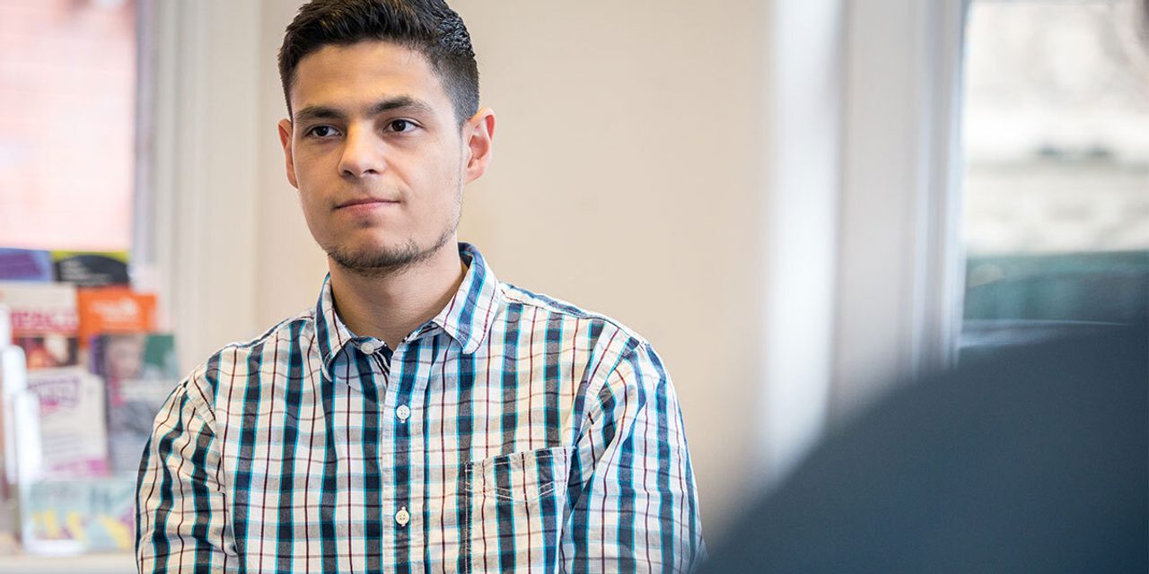 Portrait of a person with short dark hair and light stubble in a checked shirt, sitting in an office with leaflet racks behind.