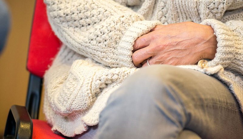 Close-up of a person's hands resting on their lap, wearing a cream cable-knit cardigan and a ring, on a red chair.