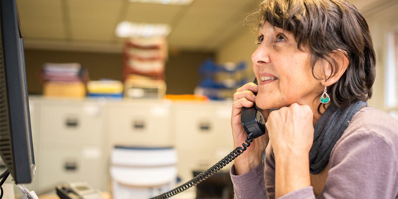 A person with short dark hair and a teardrop earring smiling while talking on a desk phone in an office with filing cabinets.
