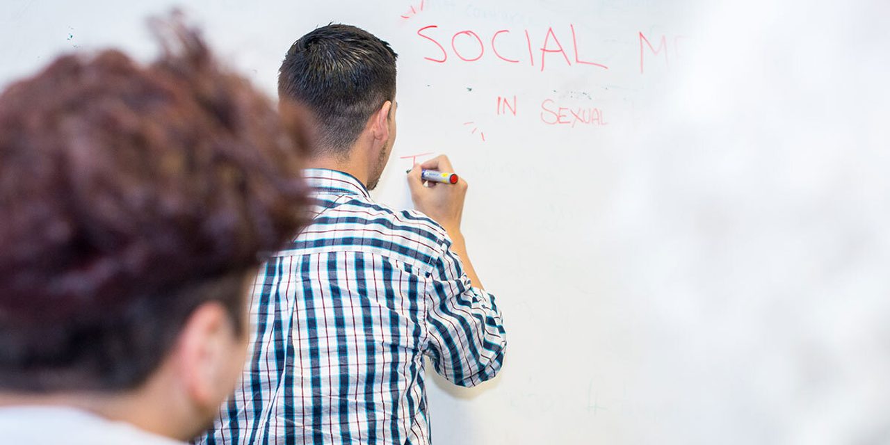 A person in a checked shirt writing "SOCIAL MEDIA IN SEXUAL..." on a whiteboard while another person watches from the foreground.