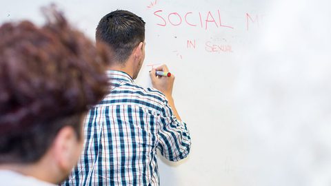 A person in a checked shirt writing "SOCIAL MEDIA IN SEXUAL..." on a whiteboard while another person watches from the foreground.