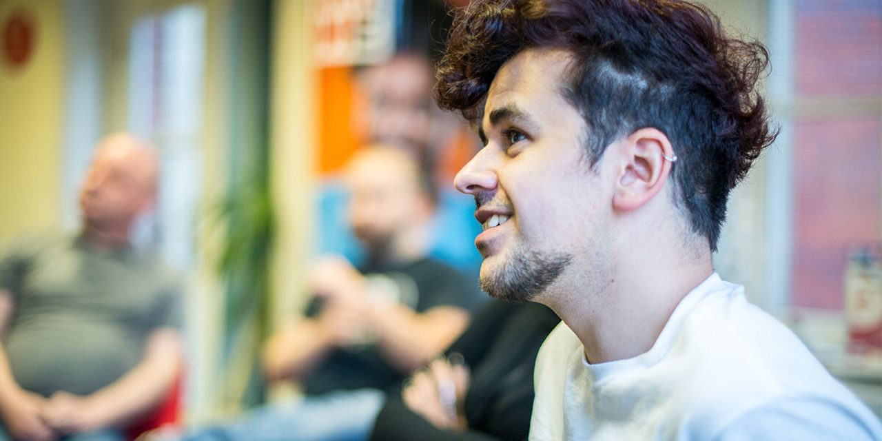 Profile portrait of a person with curly dark hair and an ear piercing, smiling during a group discussion, with others blurred behind.