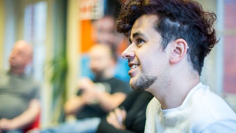 Profile portrait of a person with curly dark hair and an ear piercing, smiling during a group discussion, with others blurred behind.