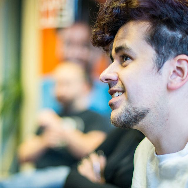 Profile portrait of a person with curly dark hair and an ear piercing, smiling during a group discussion, with others blurred behind.
