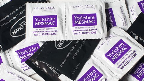 A pile of Yorkshire MESMAC condom sachets and black water-based lube sachets.
