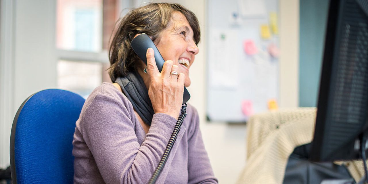A person in a purple top laughing while talking on a desk phone, sitting on a blue office chair.