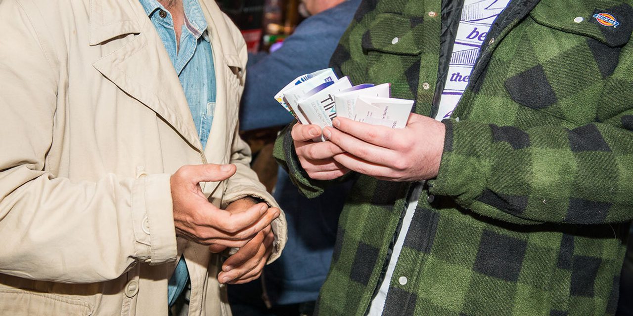 Close-up of two people outdoors at night; one in a green checked jacket holding leaflets, handing one to another in a beige coat.