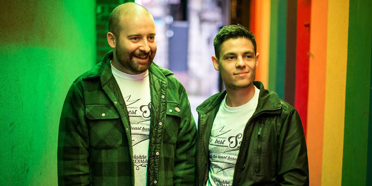 Two people standing together in an alley lit with rainbow-coloured light, wearing matching Yorkshire MESMAC t-shirts.