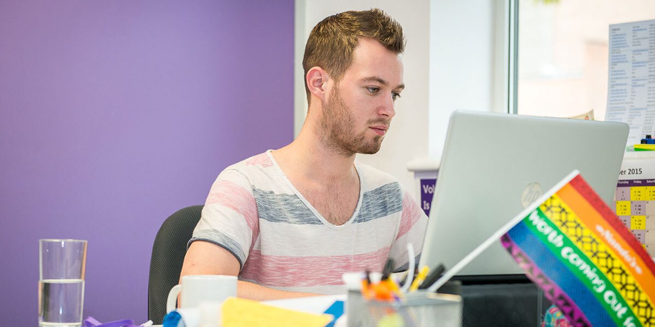 A person in a striped t-shirt working at a laptop at a desk, with a rainbow "Worth Coming Out" flag beside them, against a purple wall.