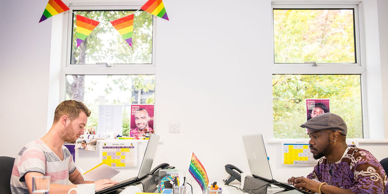 Two people working at laptops at facing desks in a bright office decorated with rainbow bunting and flags.
